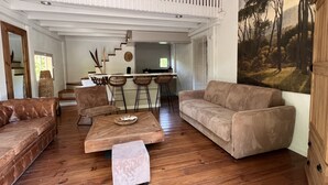A spacious living area with a wooden floor, a brown leather sofa, a matching armchair, and a small ottoman. A wooden coffee table is in the center. A kitchen area with a bar and stools is visible in the background. - Mas D'auseu (Plan-d'Orgon)