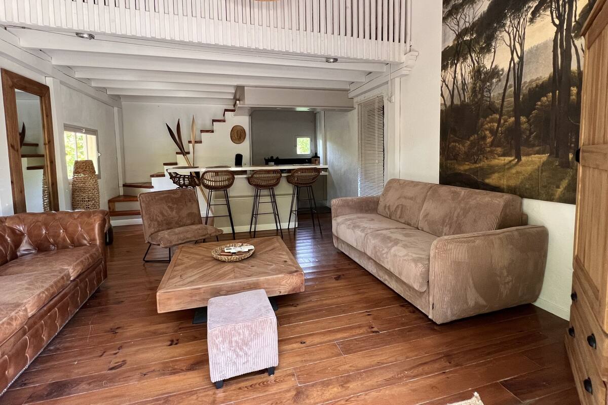 A spacious living area with a wooden floor, a brown leather sofa, a matching armchair, and a small ottoman. A wooden coffee table is in the center. A kitchen area with a bar and stools is visible in the background.