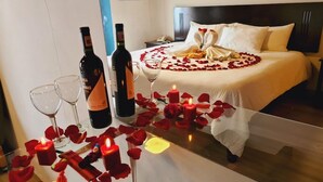 Une chambre d’hôtel avec un lit, une table de chevet ornée de bougies et de pétales de rose, et deux bouteilles de vin.