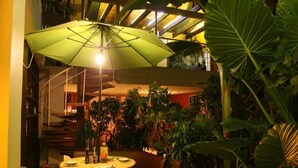 Un coin repas avec une table, des chaises et un parasol vert. On aperçoit de grandes plantes et un escalier à l’arrière-plan.