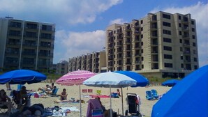 Een strand met meerdere blauwe parasols en enkele mensen die ontspannen. Achtergrond met hoge gebouwen.