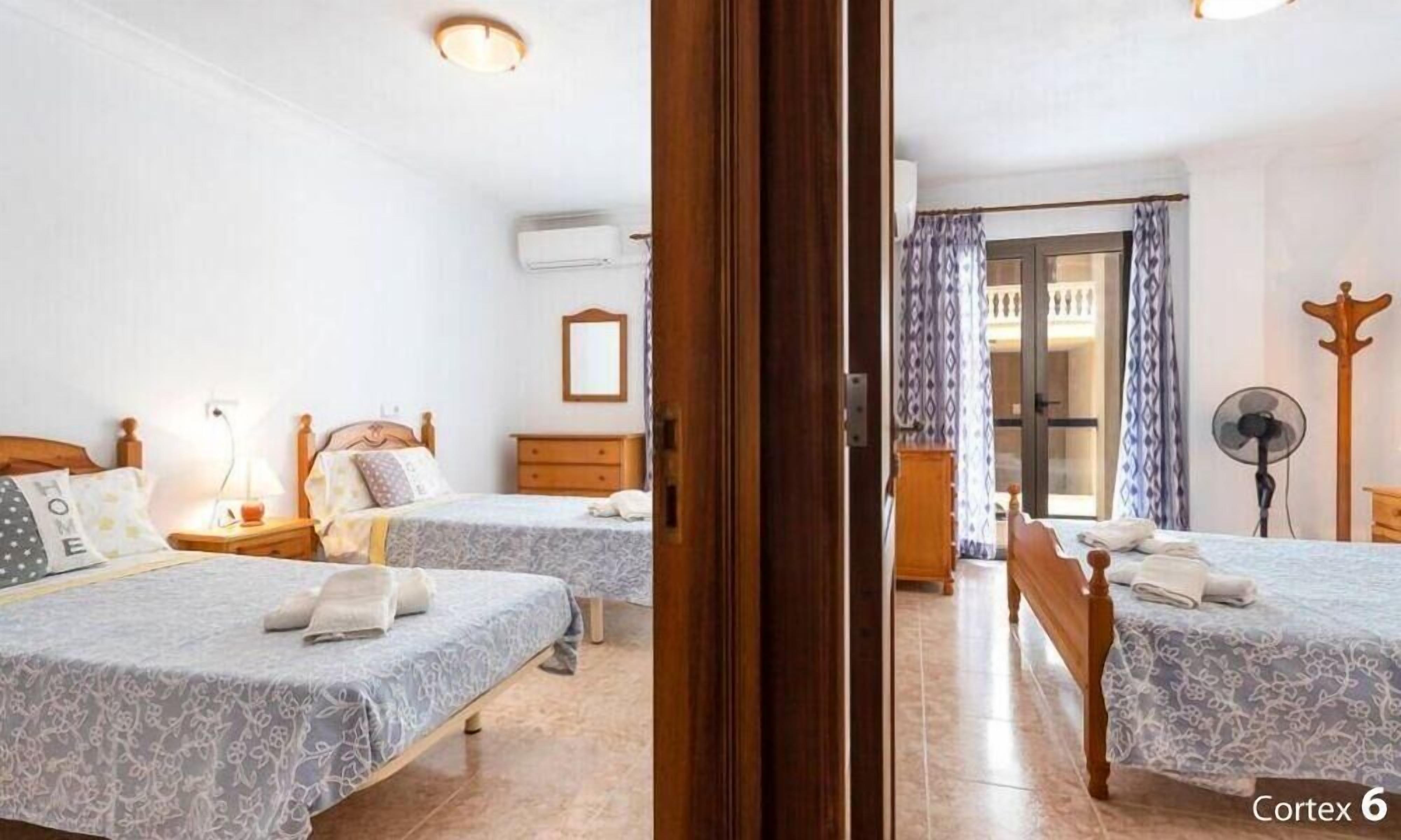 A hotel room with two beds, a wooden wardrobe, a bedside table with a lamp, and a fan.
