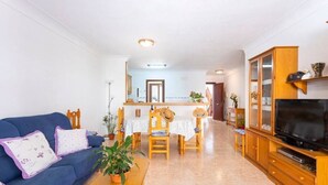 A living room with a blue sofa, wooden dining table, and chairs, a TV on a wooden cabinet, and a kitchen area in the background. - M0 Large apartment of 100 m2 + terraces (Sant Jordi)