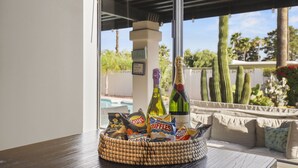 En korg av rotting med snacks och drycker står på ett bord nära ett fönster med utsikt över en pool och kaktusar.