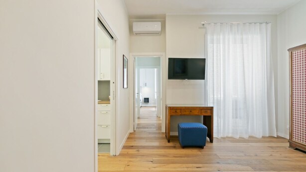 A modern living room with a wooden floor, a TV mounted on the wall, a blue ottoman, and a white curtain. - GuestHost - Elegant Don Giovanni flat of 60 sqm, for 4 people, located on the 5th floor of a historic building in Via Principe di Belmonte: NotHotel, a luxury Guest House, equipped with every comfort. Once inside you will be greeted by an elegant environ (Palermo)