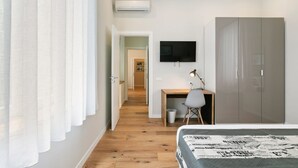 A modern bedroom with a wooden floor, a bed with a patterned duvet, a desk with a chair, a television, and a wardrobe. - GuestHost - Elegant Don Giovanni flat of 60 sqm, for 4 people, located on the 5th floor of a historic building in Via Principe di Belmonte: NotHotel, a luxury Guest House, equipped with every comfort. Once inside you will be greeted by an elegant environ (Palermo)