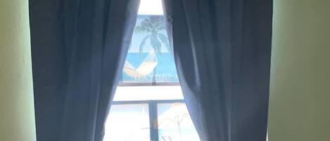 A window with dark curtains, a view of the sea, and an air conditioning unit below.