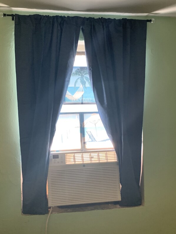 A window with dark curtains, a view of the sea, and an air conditioning unit below.