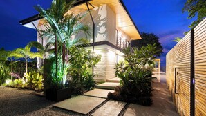 Une maison moderne dotée d’un hall d’entrée bien éclairé, entourée de plantes tropicales et bordée d’un chemin de gravier.