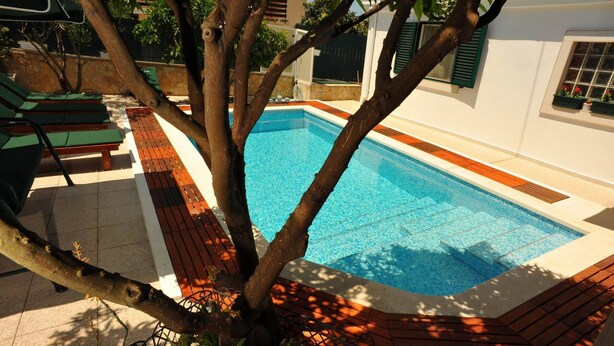 A rectangular swimming pool with a wooden deck surrounding it, a tree in the foreground, and a white building with windows and shutters in the background. - Villa Anni - Two-Bedroom Apartment with Terrace (Sutivan)