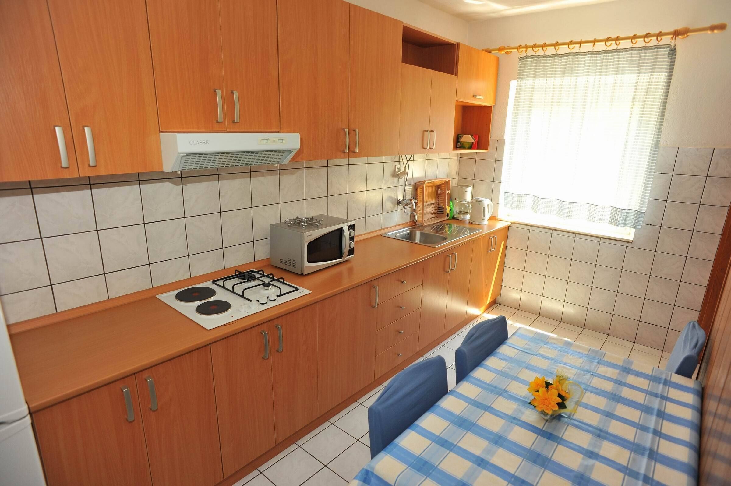 A compact kitchen with wooden cabinets, a gas stove, a microwave, and a sink. There's a window with a curtain, and a small table with a blue and white checkered tablecloth and a vase of flowers.