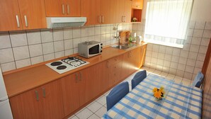 A compact kitchen with wooden cabinets, a gas stove, a microwave, and a sink. There's a window with a curtain, and a small table with a blue and white checkered tablecloth and a vase of flowers. - Villa Anni-Two Bedroom Apartment with Sea View Terrace (Sutivan)