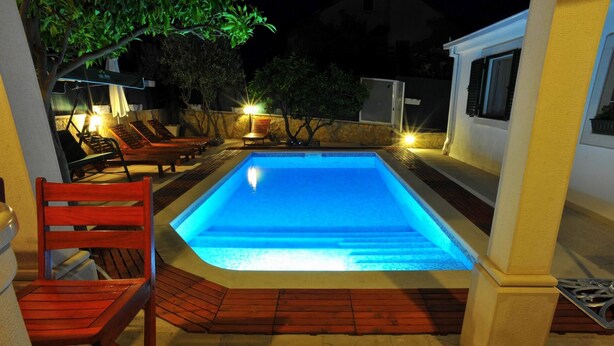 Ein Schwimmbecken mit Liegestühlen und einer Holzterrasse, umgeben von einem weißen Gebäude mit einer Steinmauer.