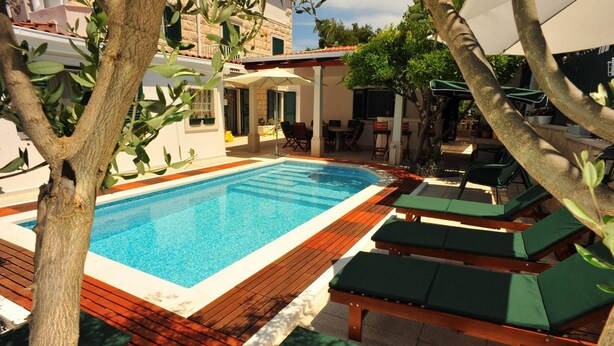Ein Schwimmbecken mit Holzterrasse und Liegestühlen, umgeben von Bäumen und Gebäuden.