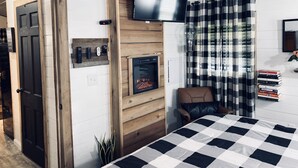 A bedroom with a checkered bedspread, a mounted TV, a fireplace, a chair, and a bookshelf with books. - Coco's Cabin #3, Where pets stay free (Goodlettsville)