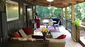 A covered porch with a wooden ceiling and a ceiling fan, a wicker sofa with cushions, a small table with a vase of flowers, and a view of a patio area with outdoor furniture. - Coco's Cabin #3, Where pets stay free (Goodlettsville)