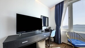A modern hotel room with a large flat-screen TV, a desk, a chair, and a view of the sea through a window. - Hotel HYGGELIG - Hostel (Busan)