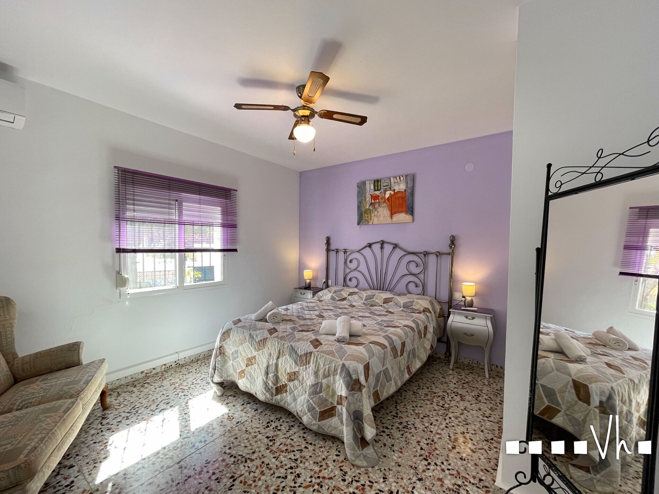Un dormitorio con cama, ventilador de techo, ventana con persianas, mesita de noche y una silla.