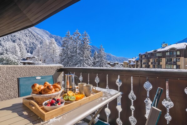 A balcony with a tray of breakfast items, including bread, fruit, and a coffee pot, overlooking a snowy mountain landscape and buildings.