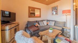 Una sala de estar acogedora con un sofá gris, techo de madera, un televisor sobre un mueble de madera, una lámpara y un cuadro enmarcado en la pared.
