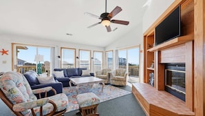A spacious living room with a ceiling fan, large windows, a fireplace, and a television. - 1509 - Sandpiper - OBX Corolla Beach House w/ Pool & Hot Tub (Corolla)