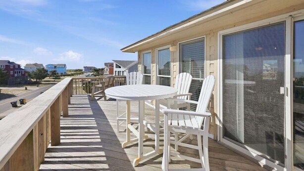A wooden deck with a table and chairs, a glass door, and a view of houses in the background. - 1509 - Sandpiper - OBX Corolla Beach House w/ Pool & Hot Tub (Corolla)
