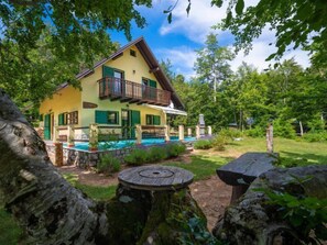 A two-story house with a balcony, a swimming pool, and a garden with trees and a wooden table.