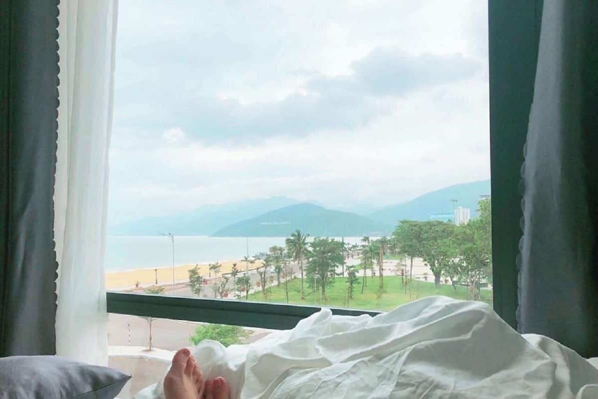 A bed with a view of a beach and mountains through a window.