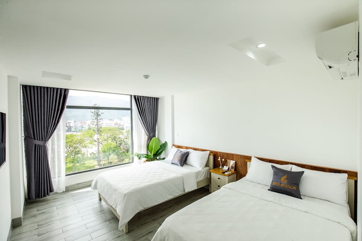 A hotel room with two beds, a large window with curtains, a wooden headboard, and a nightstand with a lamp.