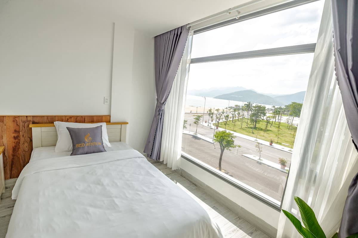 A hotel room with a large window, a bed with white bedding, a pillow with "BED STATION" text, and a view of a cityscape and mountains.