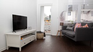 A living room with a flat-screen TV on a white TV stand, a gray sofa with cushions, a small round table, and a door leading to an outdoor balcony.