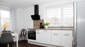 A modern kitchen with white cabinets, a black range hood, a window with blinds, a dining area with a chair, and a refrigerator. - Ferienwohnung West - Ferienwohnungen Heimathafen (Edertal)