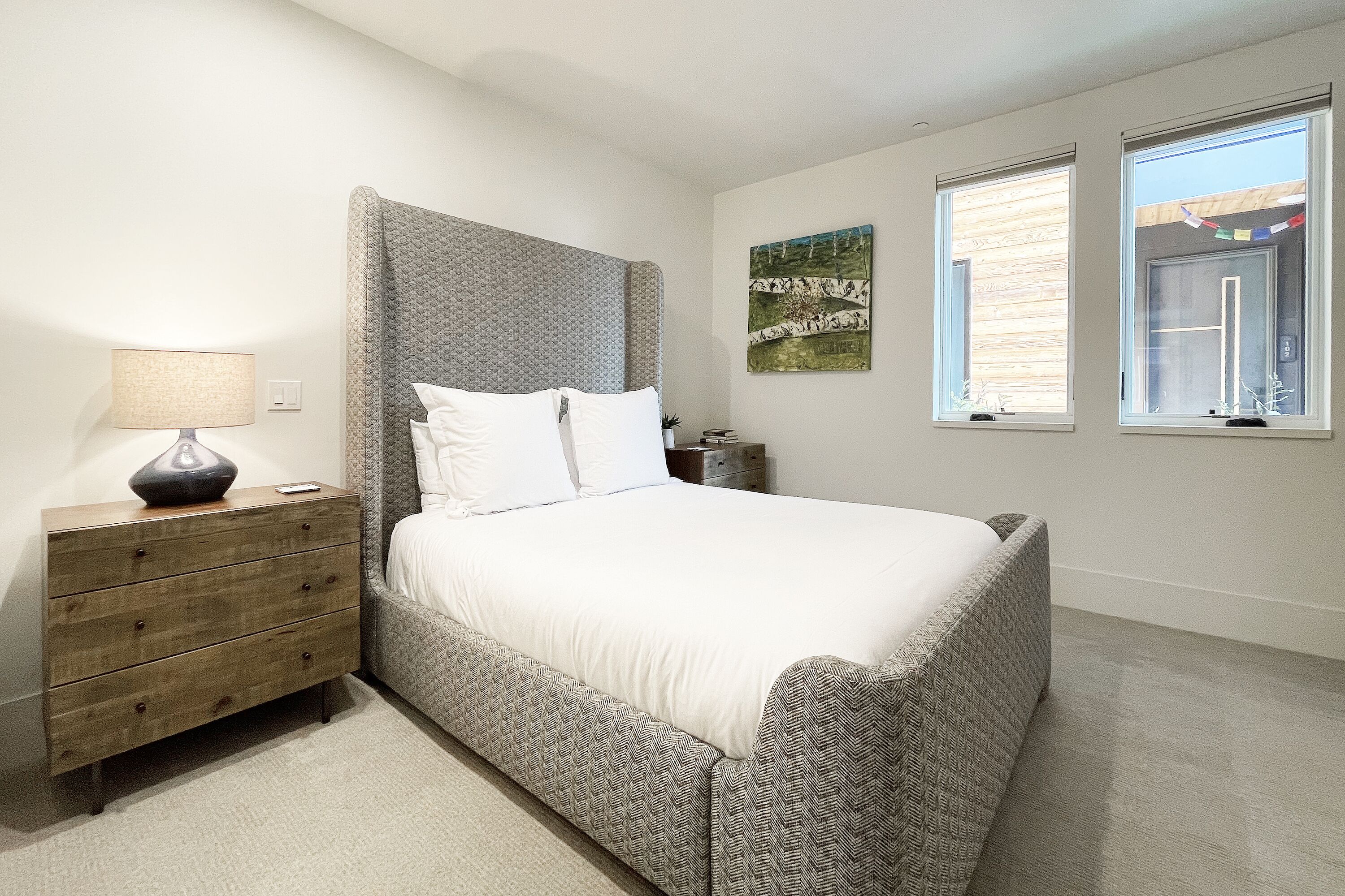 A bedroom with a bed, a dresser, a lamp, and two windows.