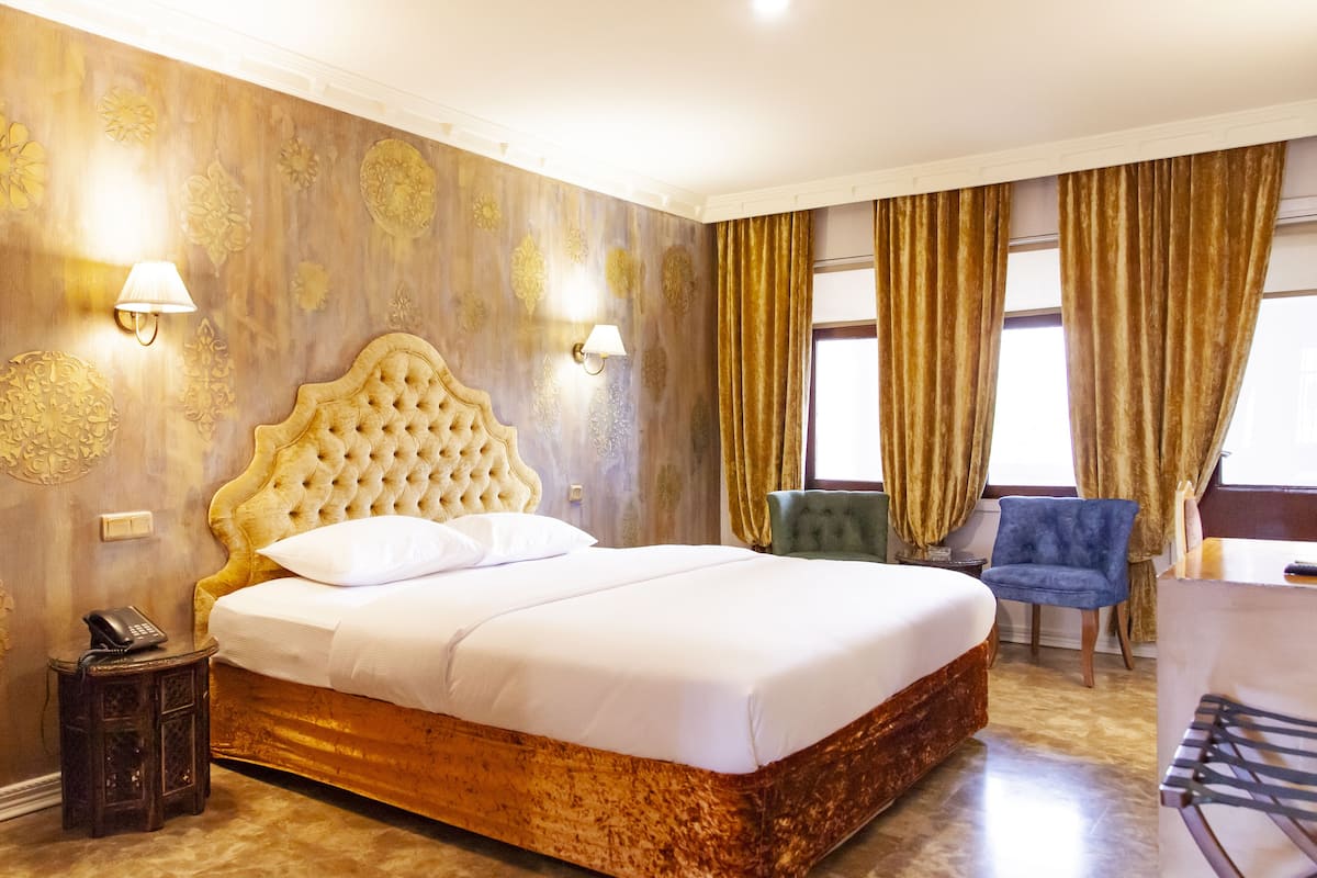 A large bed with a tufted headboard, two bedside lamps, a telephone on a side table, and a chair by the window.