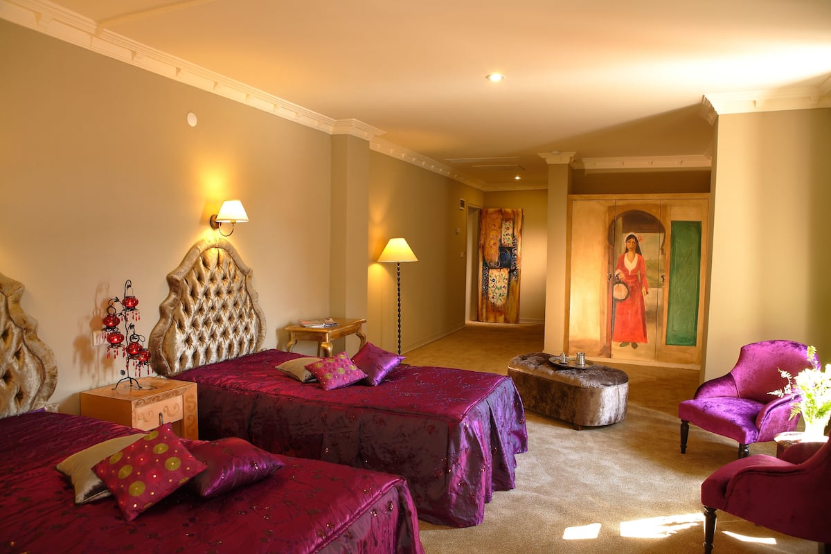 A hotel room with two beds, a chair, a lamp, and a painting on the wall.