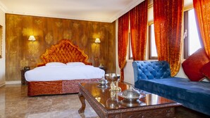 A hotel room with a large bed, a blue sofa, a wooden coffee table, and a red accent pillow. - Ala Han Boutique Hotel (Sanliurfa)