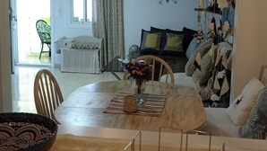 Un coin repas avec une table dressée pour deux personnes, un luminaire suspendu et une vue sur un salon agrémenté d’un canapé et d’objets décoratifs.