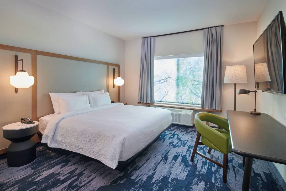 Une chambre d’hôtel avec un grand lit, un bureau, une chaise, une table de chevet, une lampe et une fenêtre avec des rideaux.