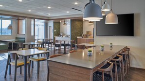 A modern dining area with a long communal table, several smaller tables, and bar stools. There is a kitchen area in the background, a large flat-screen TV mounted on the wall, and a window providing natural light.