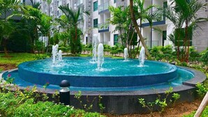 A circular pool with multiple fountains surrounded by lush greenery and a modern building in the background. - Arcadia beach condo by Jiabo (Pattaya)