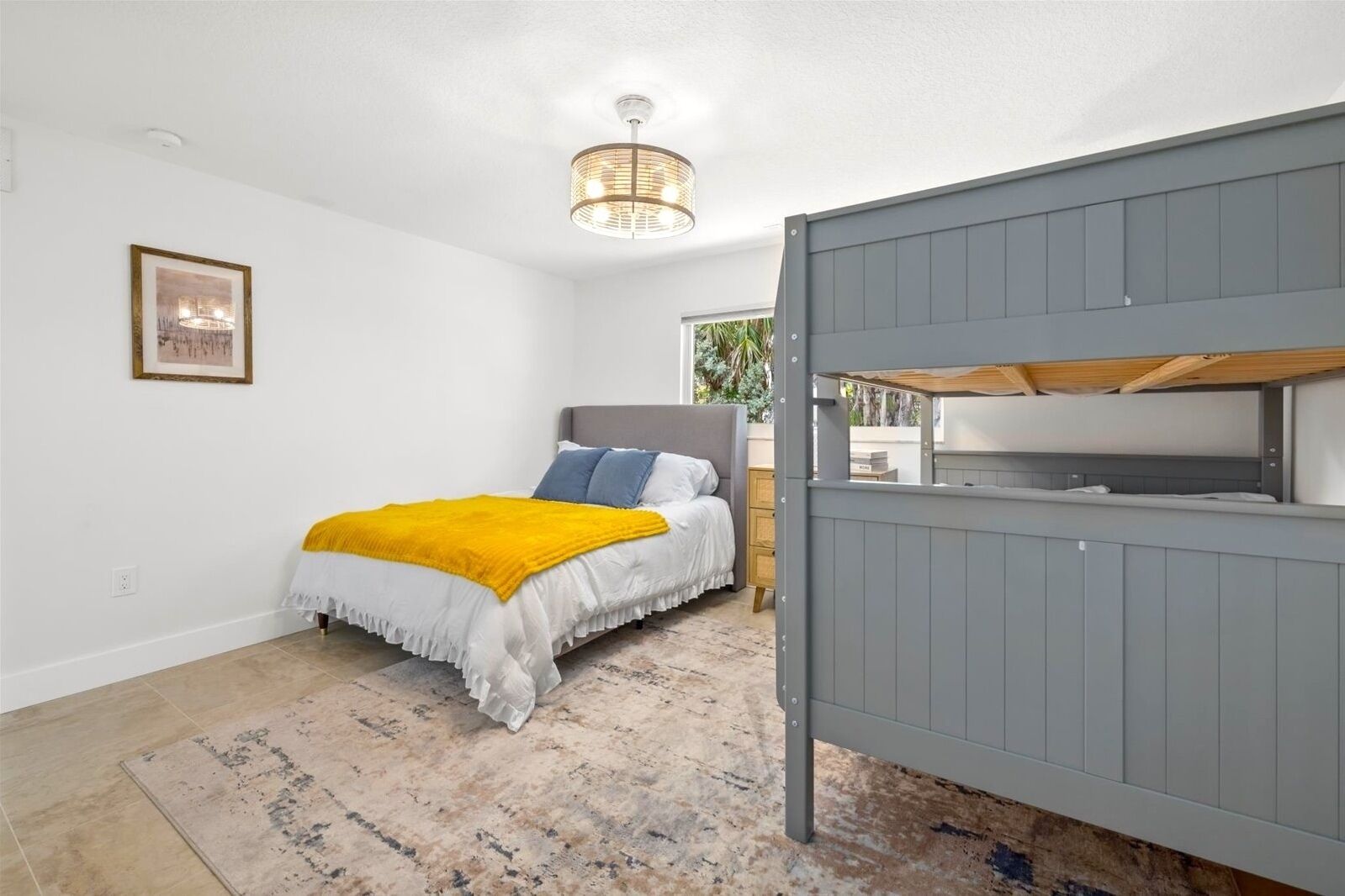 A bedroom with a bed, a grey bunk bed, a yellow throw blanket, a framed picture on the wall, and a ceiling light fixture.