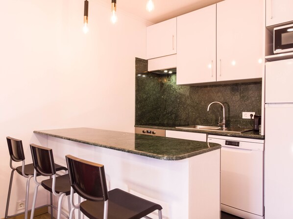 A modern kitchen with a marble countertop, stainless steel sink, and a dishwasher. There are three black chairs at the bar.