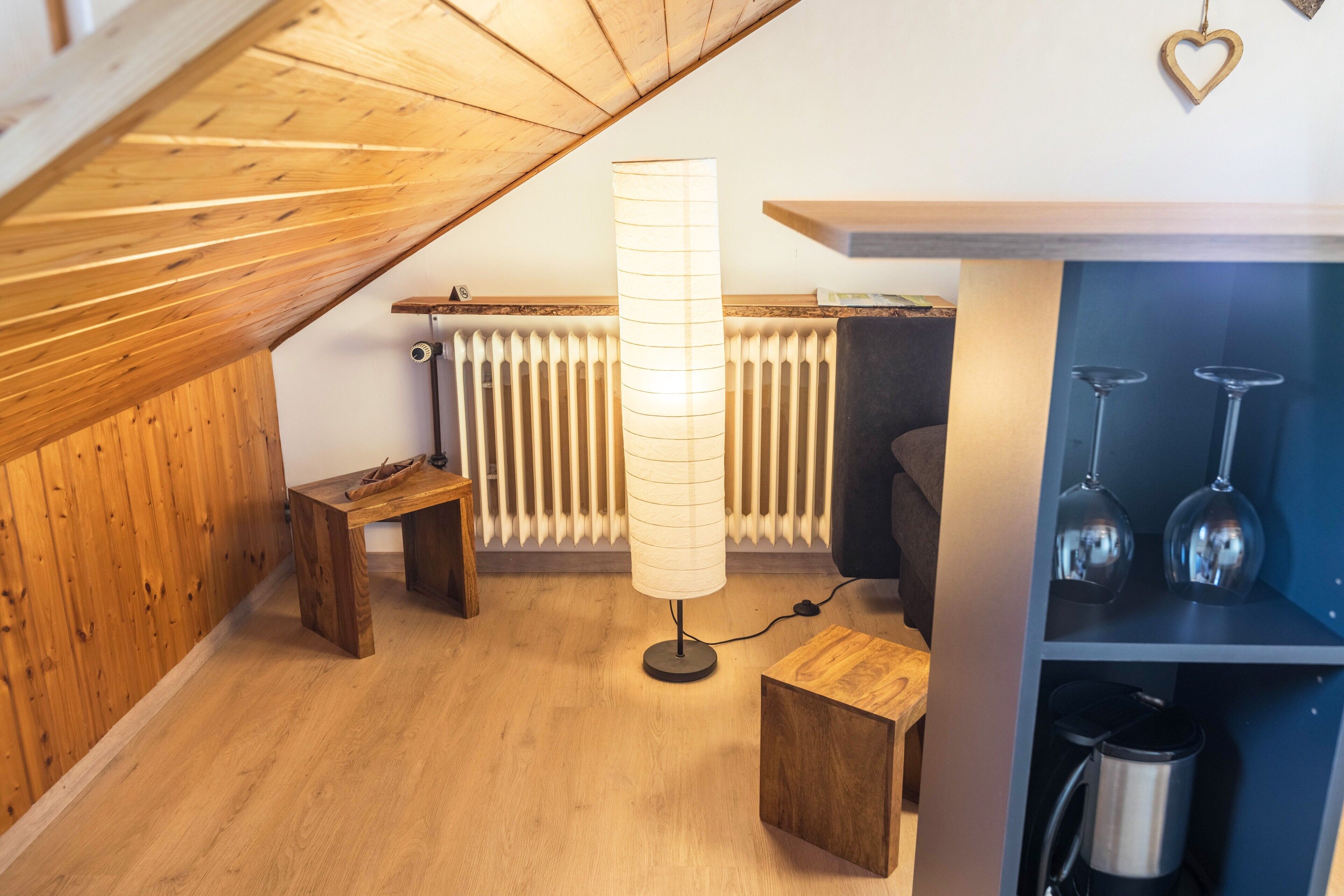 Ett rum i loftstil med ett sluttande tak, en golvlampa, en träbänk, ett litet bord, ett element och en hylla med vinglas och en termos.