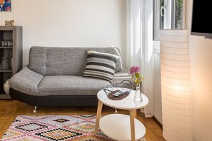 Una sala de estar moderna con un sofá gris, una mesita blanca con una cesta, un tapete estampado y una ventana con persianas.