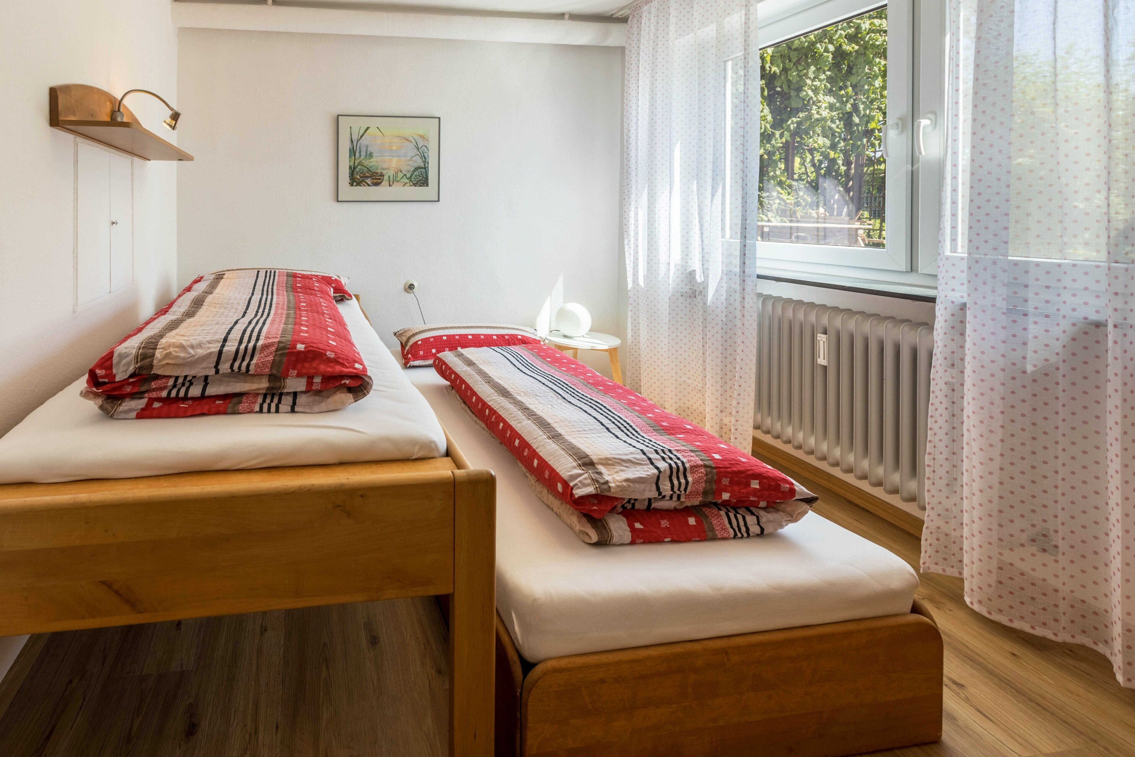 A room with two wooden beds, a small white table, a wall-mounted lamp, a framed picture, and a window with curtains.
