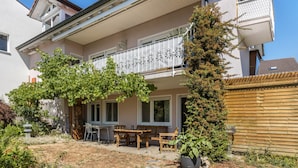 Une maison à deux étages dotée d’un balcon, d’une terrasse avec des meubles d’extérieur et d’un jardin où poussent différentes espèces de plantes.