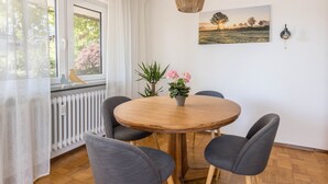Une table ronde en bois, entourée de quatre chaises grises, un pot de fleurs avec des fleurs roses, un radiateur, une fenêtre avec des rideaux blancs et un luminaire fixé au mur.