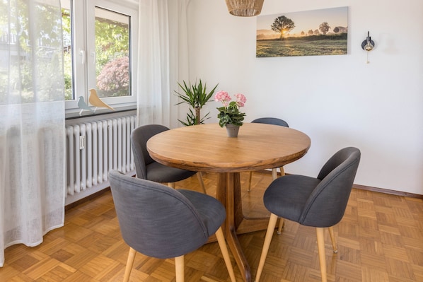 Une table ronde en bois, entourée de quatre chaises grises, un pot de fleurs avec des fleurs roses, un radiateur, une fenêtre avec des rideaux blancs et un luminaire fixé au mur.