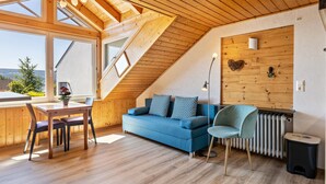 A loft-style room with a wooden ceiling, a blue sofa, a dining table with chairs, and a view of the outdoors. - Cozy Holiday Apartment "Sonnenuntergang" on Lake Constance with Lake View & Wi-Fi (Gaienhofen)