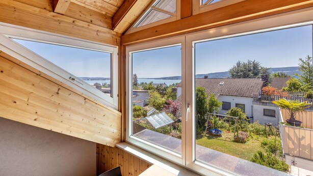 A room with a sloped ceiling, skylights, and large windows offering a view of a garden and neighboring buildings. - Cozy Holiday Apartment "Sonnenuntergang" on Lake Constance with Lake View & Wi-Fi (Gaienhofen)